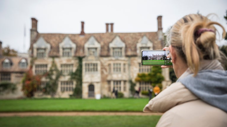 A lady takes a photo of the Anglesey Abbey house on her phone. The phone is in focus.